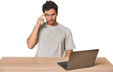 Hispanic young man working on laptop pointing temple with finger, thinking, focused on a task.