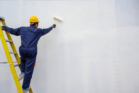 A painter holds a paint roller and skillfully paints a house. A house under construction nearing completion has a painter applying a white primer.