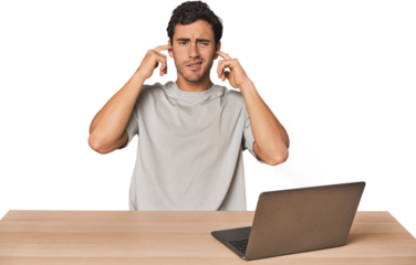 Hispanic young man working on laptop covering ears with fingers, stressed and desperate by a loudly ambient.