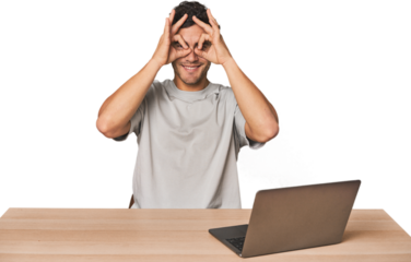 Hispanic young man working on laptop showing okay sign over eyes