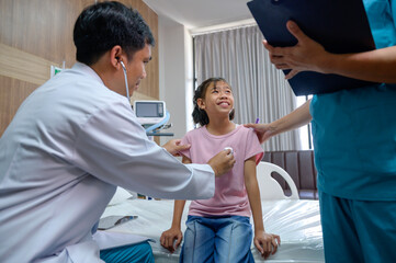 Asian male doctor using stethoscope to check pulse of girl in hospital examining and treating child patient with nurse beside him