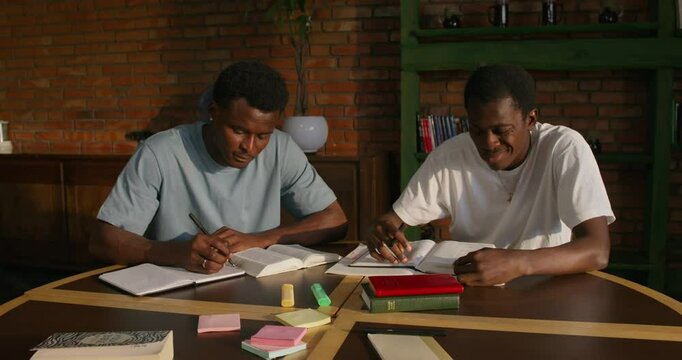 Two men study the Bible together. They are sitting at a table indoors. One of them is writing in a notebook