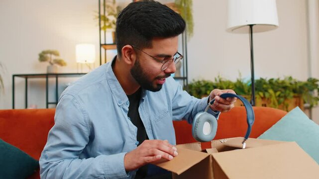 Dissatisfied annoyed Indian male customer open cardboard box receive damaged broken wireless headphones, shocked young man consumer having problem complaint frustrated with bad order delivery at home.