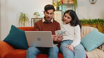 Happy young Indian couple counting money and booking tickets for holiday vacation sits on sofa in living room at home. Diverse family holding banknotes planning online investment budget home finance - Powered by Adobe