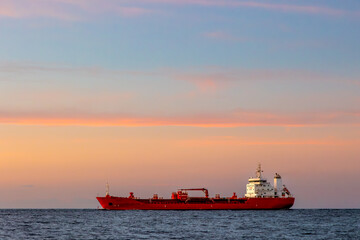 Fototapeta premium Red chemical cargo bulk ship with 'no smoking' signs heading out to flat sea or ocean under bright colourful pink and orange sunset sky