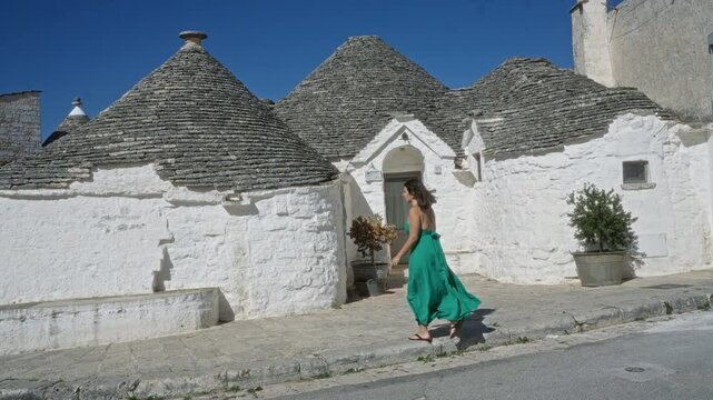 A young hispanic woman in a green dress walking along the charming trulli houses in the old town of alberobello, italy, under a clear blue sky.