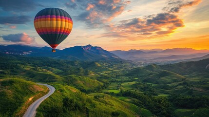 Naklejka premium Brightly colored hot air balloon flying over a serene green valley with a winding road and majestic mountains at sunrise.