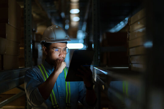 A man in a blue shirt and safety vest is looking at a tablet. He is wearing a hard hat and safety glasses