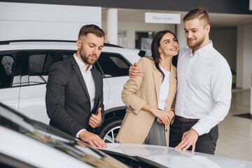 A well-dressed salesman is presenting information to couple in a car dealership showroom. The couple appears interested as they stand between two new vehicles