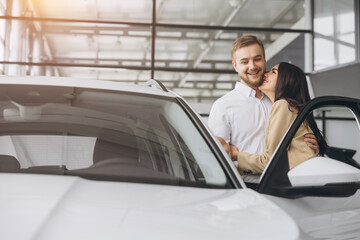 Happy young lovely couple in casual wear hugging while buying first new family car together in dealership. Man shows car keys in showroom.