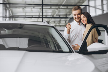 Happy young lovely couple in casual wear hugging while buying first new family car together in dealership. Man shows car keys in showroom.