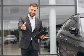 Portrait, smiling man extending hand for handshake at camera at outside car dealership for car sale...