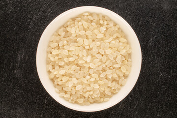 Round grain uncooked rice with white ceramic saucer on slate stone, macro, top view.