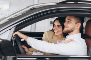 Portrait of smiling beautiful couple sitting in modern luxury car, happy excited young man and woman buying new car in dealership.