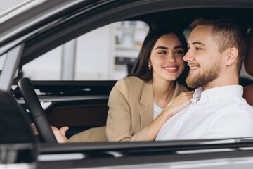 Portrait of smiling beautiful couple sitting in modern luxury car, happy excited young man and woman buying new car in dealership.