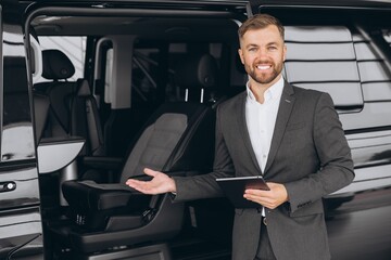 Smiling friendly car seller in suit standing in car salon and holding tablet. It's always pleasure to buy a car on a right place.