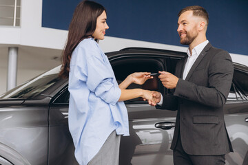 Smiling salesman or manager giving car keys and shakes hand to modern happy woman new car owner in car dealership