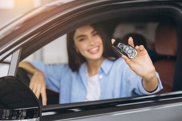 Close-up shot of happy smiling woman sitting in new car and holding keys in car dealership showroom