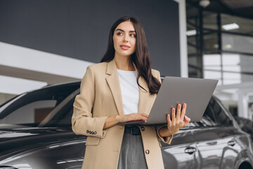 Charming car seller, businesswoman in beige suit standing in car salon and using digital laptop...