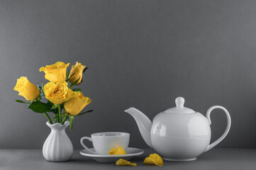 Yellow roses in a ceramic vase, white teapot and cup of tea on a gray background.
