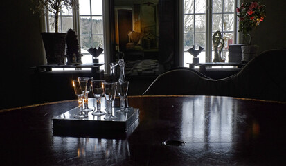 A room in darkness with a table holding fine glasses, illuminated by the light coming from the windows in the room.