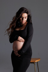 A beautiful pregnant woman with long dark hair with styling and makeup. A brunette waiting for a baby, sit on high chair in a long black dress. Grey background in the studio, fan.