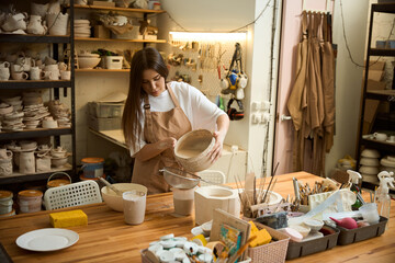 Pretty woman in apron straining the clay mixture through a sieve in creative studio