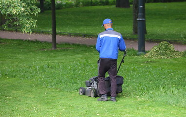 A worker in a blue uniform mows the grass with a lawn mower, Mikhailovsky Garden, St. Petersburg,...