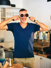A barista wearing a blue V-neck t-shirt holds a portafilter in both hands, standing behind the counter in a modern coffee shop with an espresso machine.