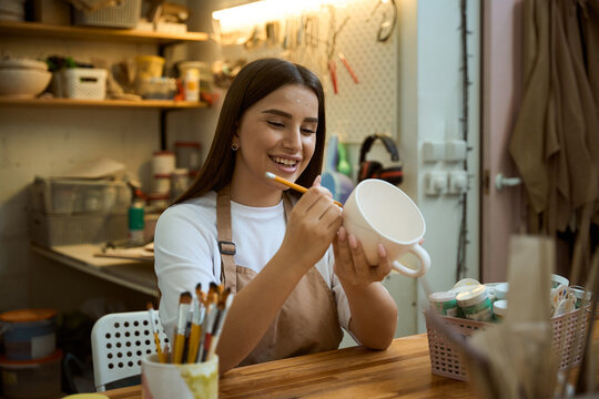 Smiling woman potter holding pencil and painting on clay cup