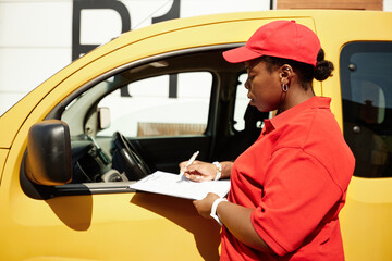Delivery person in red uniform standing beside yellow vehicle while checking and signing delivery...