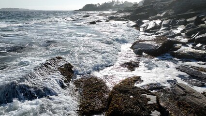 Ocean waves and surf breaking on peaceful rocky coastline  beach as sunshine reflects on water during summertime along seashore in Maine