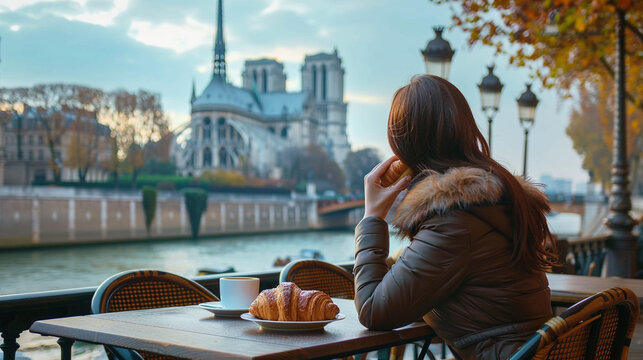 A girl sitting at a cafÃ© table, with a croissant in hand and the Notre-Dame Cathedral in the background