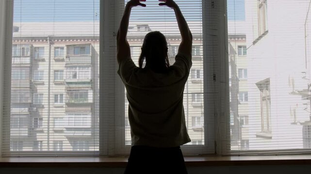 Back view of silhouette of male ballet dancer doing heel raises with arms over head, then using windowsill as barre and practicing plie while exercising in studio