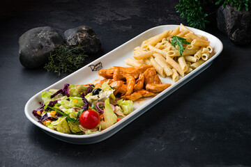 Penne pasta with fried chicken and a green salad of olives, lettuce, arugula, cherry tomatoes, corn and carrots on a dark table.