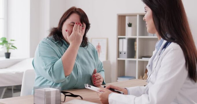 Young female doctor listening to fat patient's complaints sitting on workplace in examination room in medical clinic. Overweight woman suffering from headache. Medicine concept. 4k video.