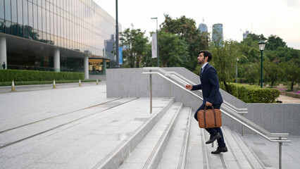 Skilled business man walking up stairs at park or city while holding bag in the hand Professional project manager going to workplace. Represent growth, getting promotion, increasing skill. Exultant.