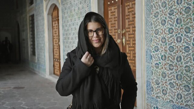 Smiling young woman with glasses and scarf standing in front of ornate tilework at topkapi palace in istanbul, turkey.