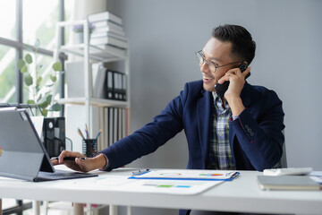 Asian businessman is working in the office, checking financial data on a digital tablet and talking on his phone