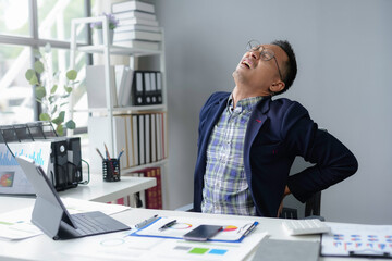 Asian office worker feeling backache sitting at his desk, he is holding his lower back with pained expression