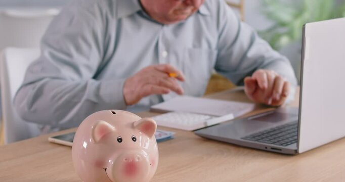 Piggy bank with a senior man working from home with financial documents in the background. Financial management, including income, pension, bills, and accounting, highlighting financial planning.