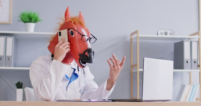 Doctor in funny silly stupid carnival masquerade horse mask, glasses, white coat and stethoscope sits at desk, talks on phone, makes agreement, uses laptop and puts consultation on his busy schedule