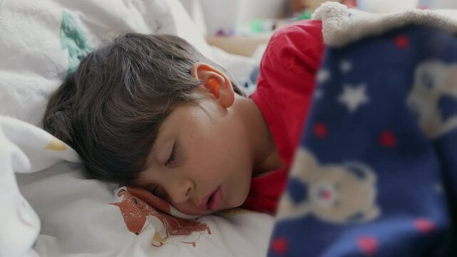 Boy sleeping peacefully under a blue blanket with teddy bear patterns, resting his head on the pillow, capturing the innocence and tranquility of childhood in a warm and cozy bed