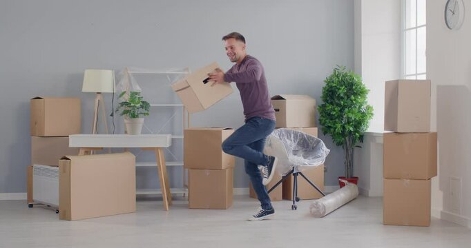 Portrait of happy excited young man carrying cardboard boxes in his new empty apartment, having fun and dancing happy to move in new house. Moving day, real estate property concept. 4k video.