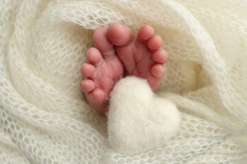 The tiny foot of a newborn baby. Soft feet of a new born in a white wool blanket. Close up of toes, heels and feet of a newborn. Knitted white heart in the legs of a baby. Macro photography. 