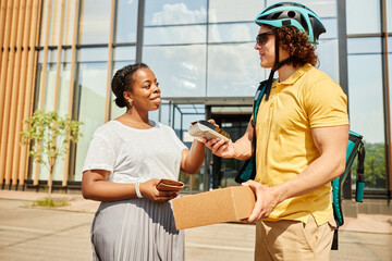 Obraz premium Smiling recipient receiving package from delivery person outside modern building. Both individuals are engaging in friendly interaction while person in helmet hands over the item