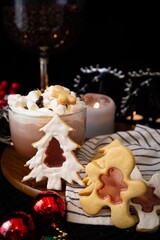 Festive hot cocoa with marshmallows, gingerbread cookies in Christmas tree shapes, on a wooden plate with holiday decorations.