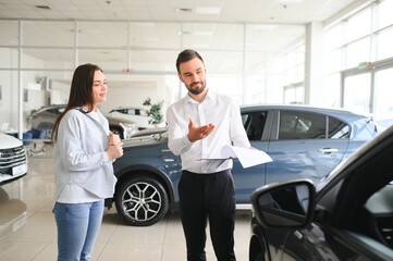 Satisfied happy caucasian female client customer woman sitting at the wheel of new car while male shop assistant helping her choose it