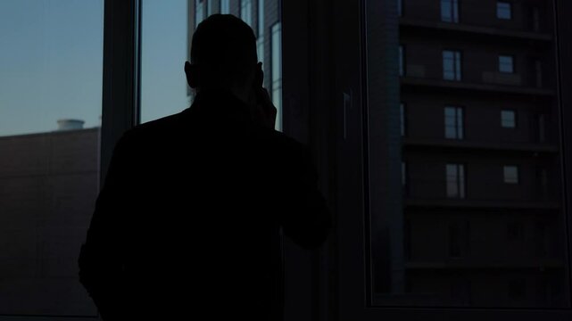 Dark silhouette of businessman entrepreneur man in office with downtown view. Mysterious man talking on phone and solving business near window. Success and motivation in financial, money and crypto