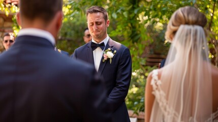 During a heartfelt outdoor wedding ceremony, the groom, dressed in a navy blue suit, expresses deep emotions as he cries while listening to the vows exchanged